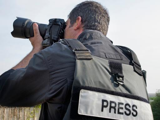 Blick auf einen Fotografen, der mit dem Rücken zum Betrachter steht. Er macht gerade mit seinen Apparat eine Aufnahme und trägt eine Sicherheitsweste mit der Aufschrift "Press". 