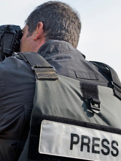 Blick auf einen Fotografen, der mit dem Rücken zum Betrachter steht. Er macht gerade mit seinen Apparat eine Aufnahme und trägt eine Sicherheitsweste mit der Aufschrift "Press". 