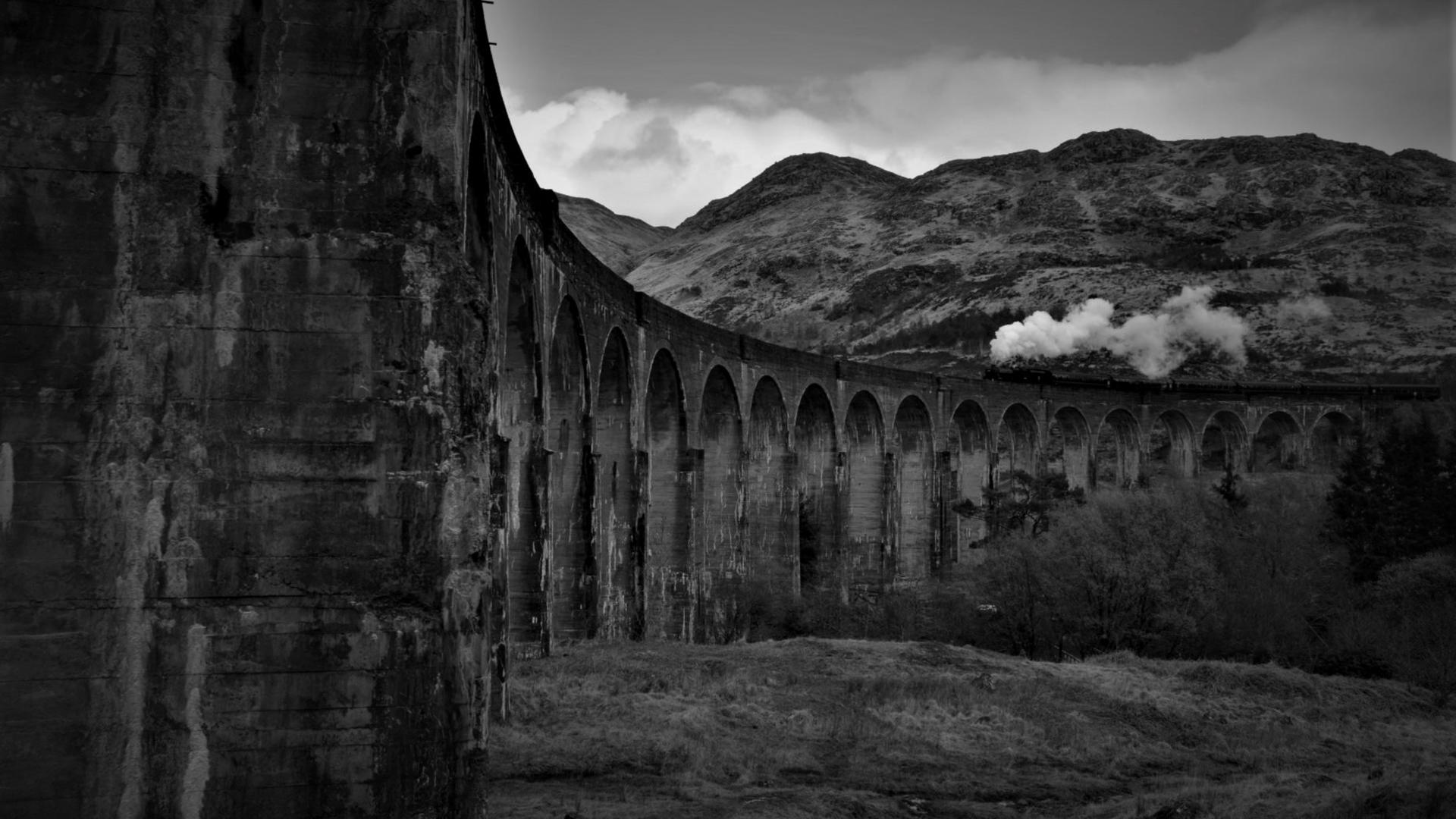 Schwarz-Weiß-Aufnahme einer Dampflok, die in der Landschaft über ein Viadukt fährt. Schwarz-Weiß-Aufnahme einer Dampflok, die in der Landschaft über ein Viadukt fährt.