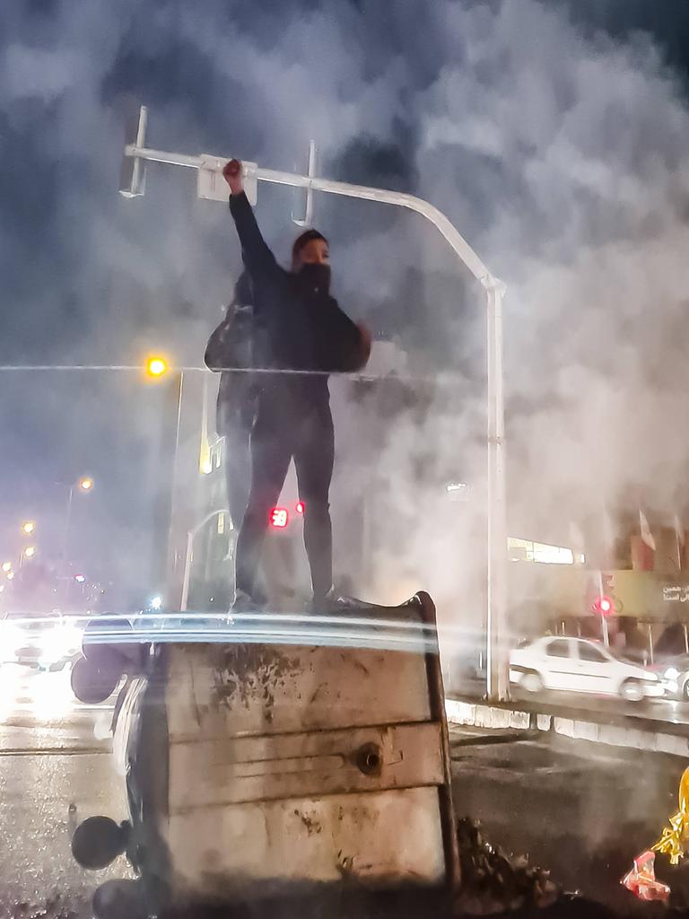 Eine Frau steht nachts auf einem umgeworfenen Müllcontainer und reckt die Faust nach oben. Um sie herum ist viel Rauch.