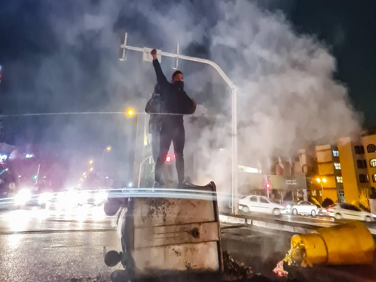Eine Frau steht nachts auf einem umgeworfenen Müllcontainer und reckt die Faust nach oben. Um sie herum ist viel Rauch.