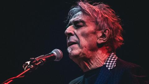 Das Foto zeigt John Cale im Profil auf der Bühne. Er trägt ein kariertes Hemd unter einem dunklen Pullover, die Haare sind struppelig, vor dem Kopf ein Mikroständer und ein Mikrofon. Cale hat die Augen geschlossen. Der Hintergrund ist dunkel.