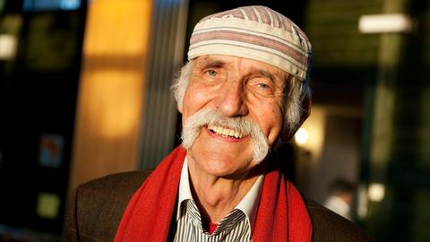 Fotografie von Ali Mitgutsch, er lächeltin der Sonne, trägt einen grauen Schnauzbart und eine weiß-braune charakteristische Mütze