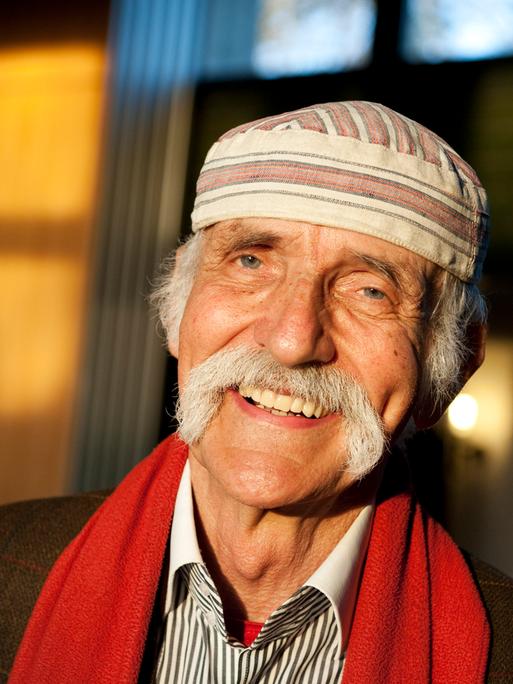 Fotografie von Ali Mitgutsch, er lächeltin der Sonne, trägt einen grauen Schnauzbart und eine weiß-braune charakteristische Mütze