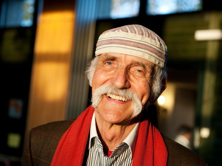 Fotografie von Ali Mitgutsch, er lächeltin der Sonne, trägt einen grauen Schnauzbart und eine weiß-braune charakteristische Mütze