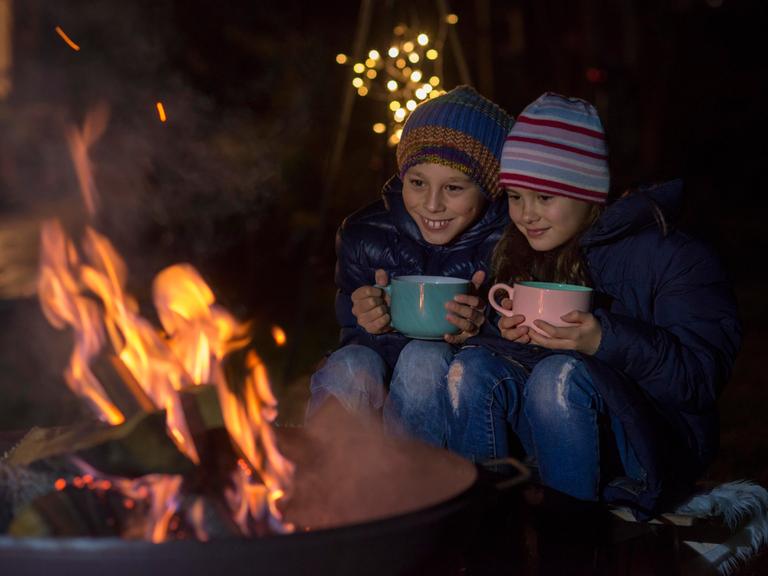 Ein Junge und ein Mädchen sitzen mit großen Tasten an einem Lagerfeuer.