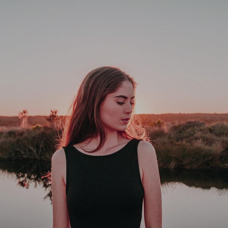 Eine junge Frau, mit dem Kopf zur Seite, steht vor einem See. Im Hintergrund ist eine flache Landschaft zu sehen.
