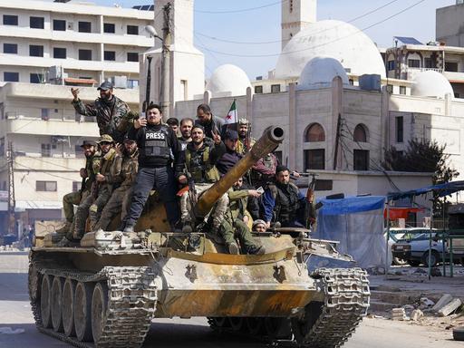 Ein Dutzend syrische Sicherheitsleute auf einem Panzer, im Hintergrund eine Moschee