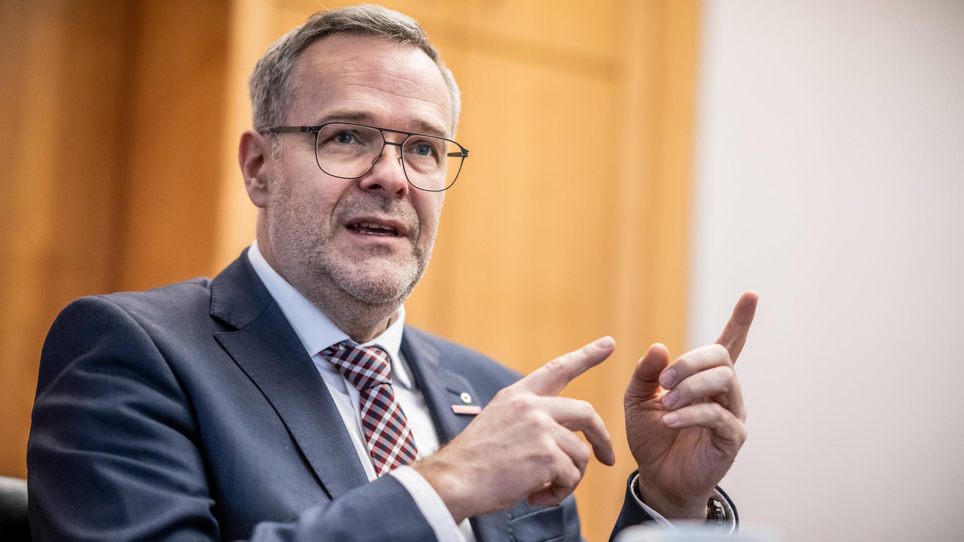 Dittrich steht in einem Raum vor einer Tür, sagt etwas und gestikuliert mit beiden Zeigefingern. Dittrich steht in einem Raum vor einer Tür, sagt etwas und gestikuliert mit beiden Zeigefingern.