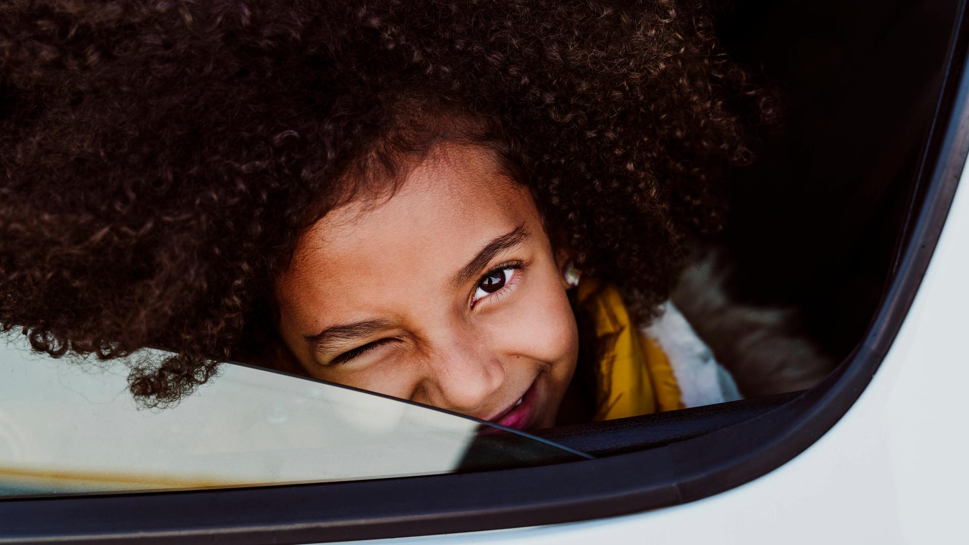 Ein Mädchen schaut mit einem zugekniffenen Auge aus einem Auto heraus und lächelt. Die Scheibe ist fast ganz heruntergelassen.