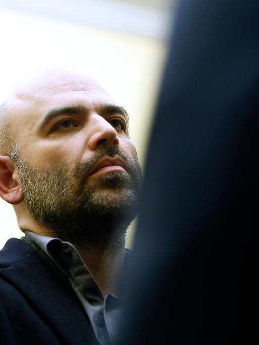 Roberto Saviano ist links im Bild zu sehen, rechts im Vordergrund ist eine dunkle Partie. Der Mittvierziger hat eine Halbglatze und trägt einen Drei-Tage-Bart.