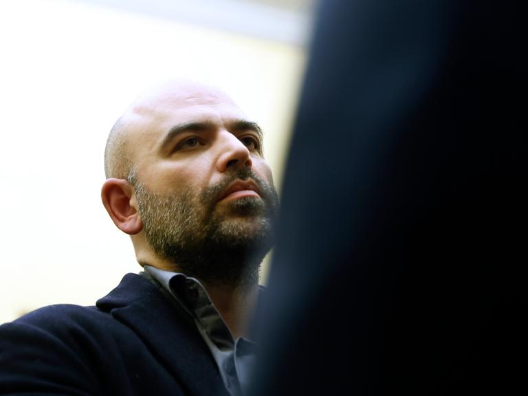 Roberto Saviano ist links im Bild zu sehen, rechts im Vordergrund ist eine dunkle Partie. Der Mittvierziger hat eine Halbglatze und trägt einen Drei-Tage-Bart.