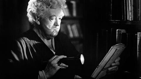 Die Schauspielerin Margaret Rutherford in ihrer Paraderolle als schrullige Miss Marple. Auf der Schwarz-Weiß-Fotografie leuchtet sie in dunkler Umgebung mit einer Taschenlampe auf ein Buch, das sie in der anderen Hand hält. 