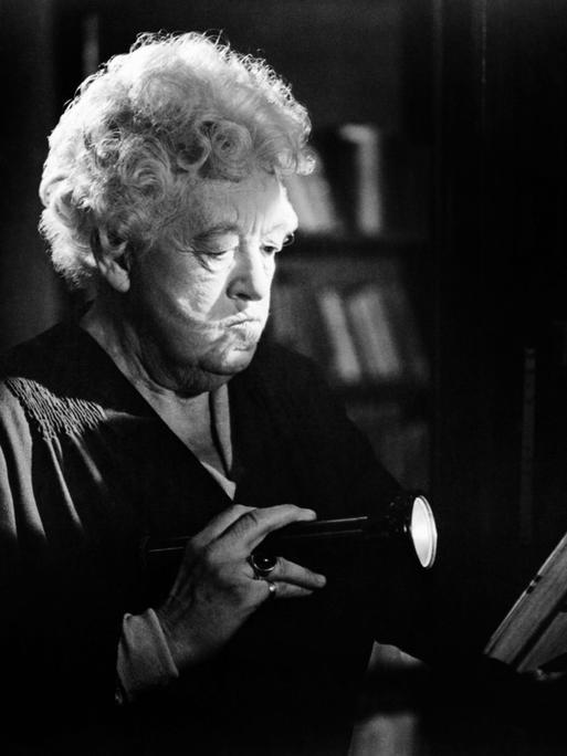 Die Schauspielerin Margaret Rutherford in ihrer Paraderolle als schrullige Miss Marple. Auf der Schwarz-Weiß-Fotografie leuchtet sie in dunkler Umgebung mit einer Taschenlampe auf ein Buch, das sie in der anderen Hand hält. 