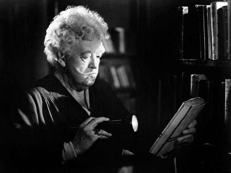 Die Schauspielerin Margaret Rutherford in ihrer Paraderolle als schrullige Miss Marple. Auf der Schwarz-Weiß-Fotografie leuchtet sie in dunkler Umgebung mit einer Taschenlampe auf ein Buch, das sie in der anderen Hand hält. 