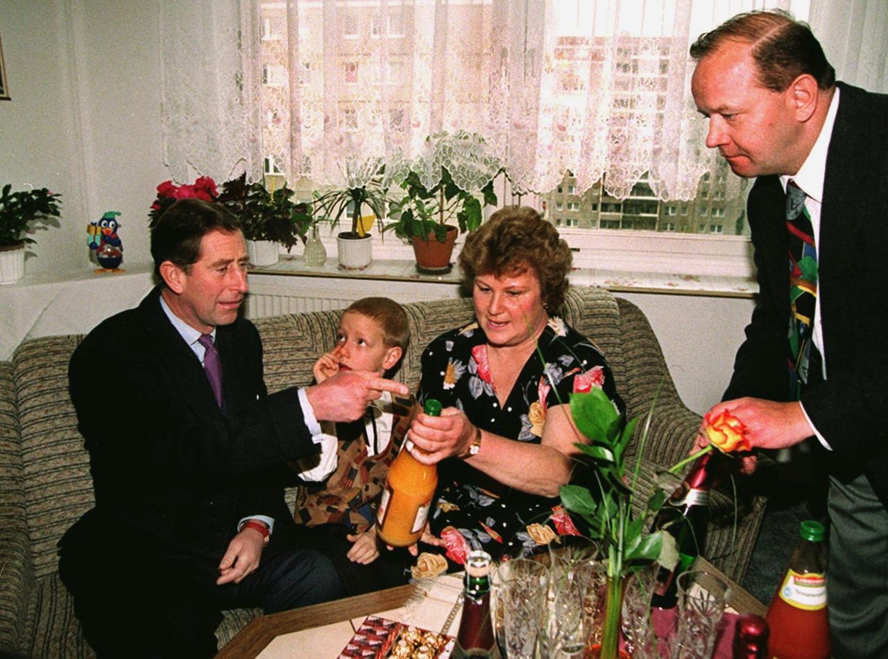 Prinz Charles sitzt mit einer Familie auf der Couch im Wohzimmer einer Plattenbau-Wohnung. 