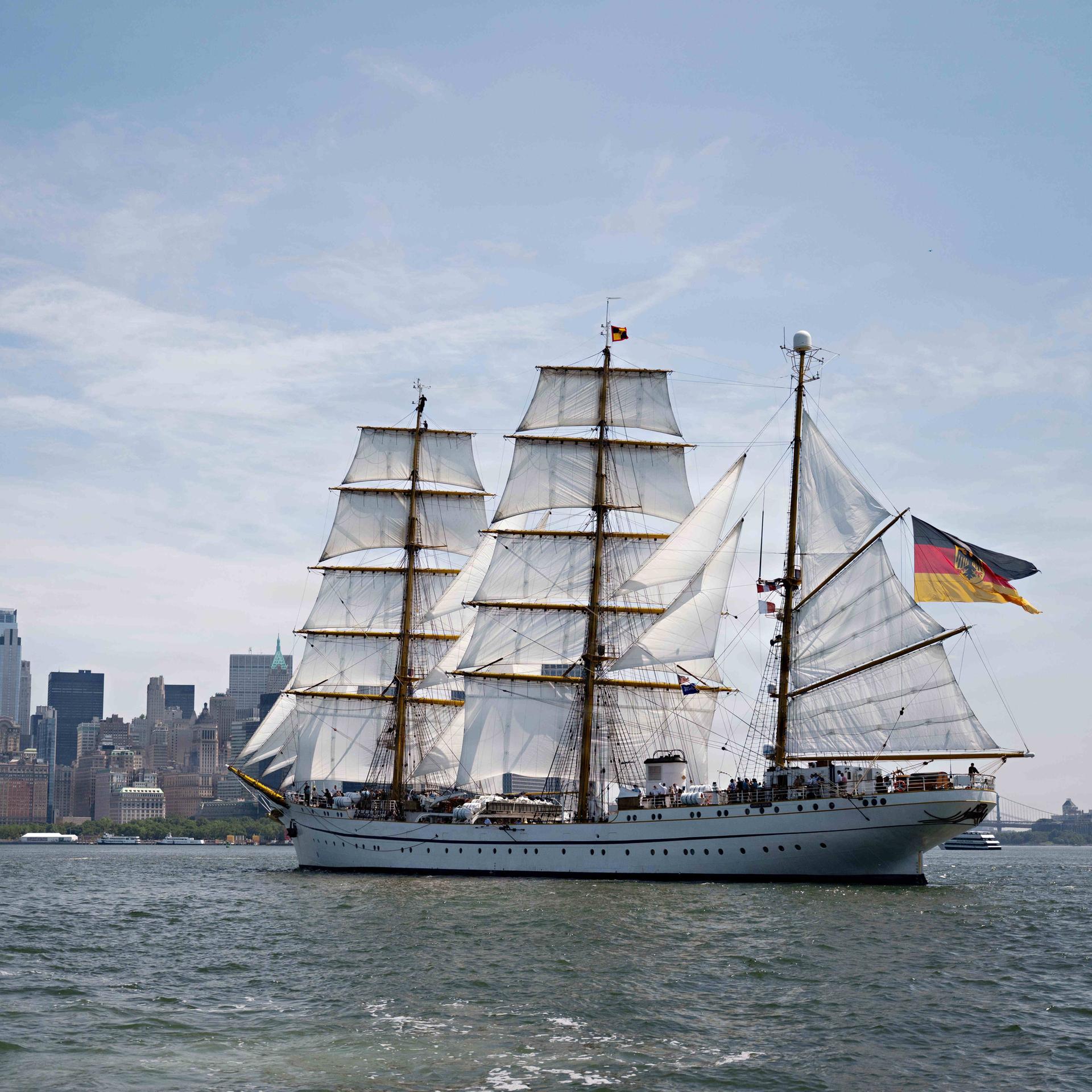 Abgebildet ist das deutsche Marineschiff „Gorch Fock“ auf dem Hudson River mit gehissten Segeln. Im Hintergrund sind die Wolkenkratzer Manhattans. 