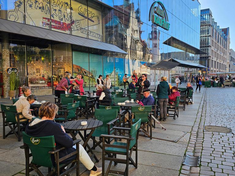 Menschen sitzen im Freien auf dem Marktplatz von Chemnitz in einem Café an Tischen.