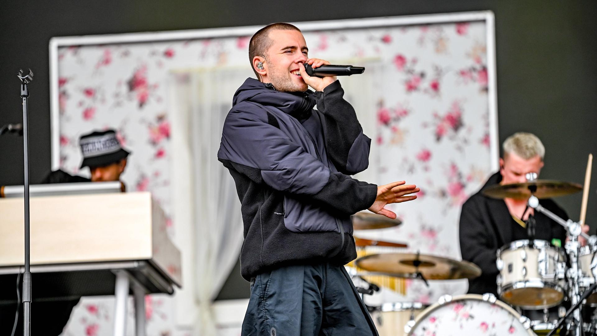 Ein Mann mit kurzgeschorenem Haar steht auf einer Bühne und singt in ein Mikrofon. Rechts neben ihm sitzt ein Schlagzeuger an seinem Instrument, links ein Keyboarder. Ein Mann mit kurzgeschorenem Haar steht auf einer Bühne und singt in ein Mikrofon. Rechts neben ihm sitzt ein Schlagzeuger an seinem Instrument, links ein Keyboarder.