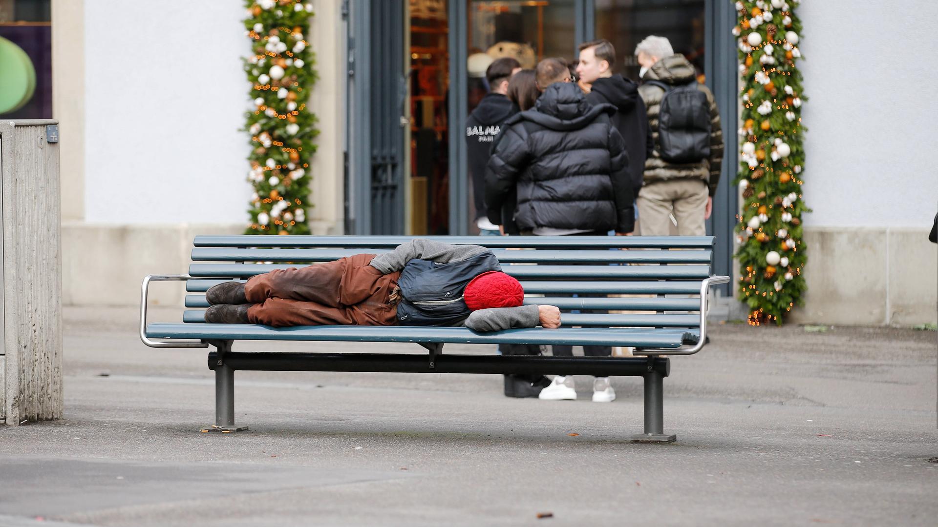 Foto Manuel Geisser 30.12.2021 Zürich : Armut . Mann auf einer Parkbank . Hinten stehen Passanten Schlange vor einem Kle