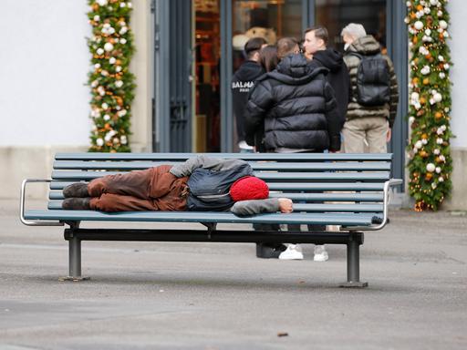 Foto Manuel Geisser 30.12.2021 Zürich : Armut . Mann auf einer Parkbank . Hinten stehen Passanten Schlange vor einem Kle