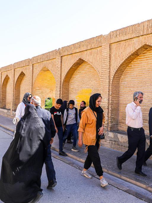 In der iranischen Stadt Isfahan laufen mehrere Passanten auf einer Straße entlang, neben der Straße steht eine Mauer mit Bögen.