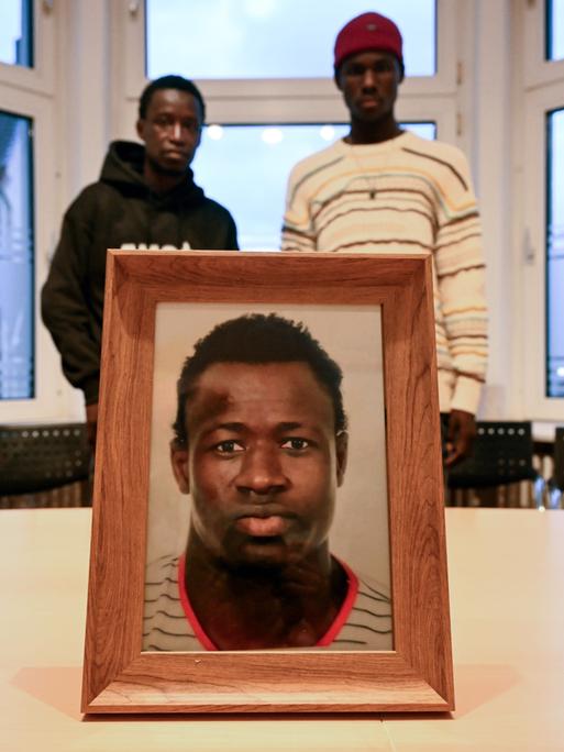 Ein gerahmtes Foto steht in einem Büroraum auf einem Tisch. Darauf ist Mouhamed Dramé zu sehen. Im Hintergrund stehen Sidy und Lassana Dramé, die Brüder des getöteten Mouhamed.