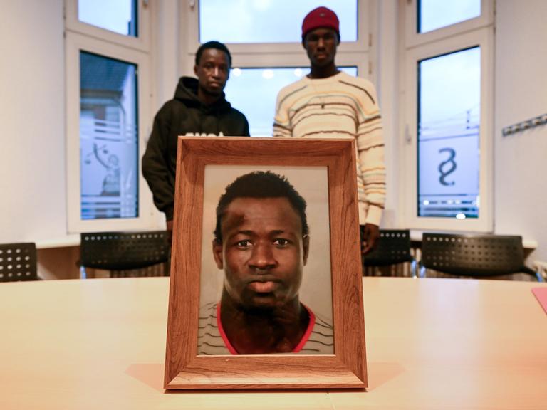 Ein gerahmtes Foto steht in einem Büroraum auf einem Tisch. Darauf ist Mouhamed Dramé zu sehen. Im Hintergrund stehen Sidy und Lassana Dramé, die Brüder des getöteten Mouhamed.