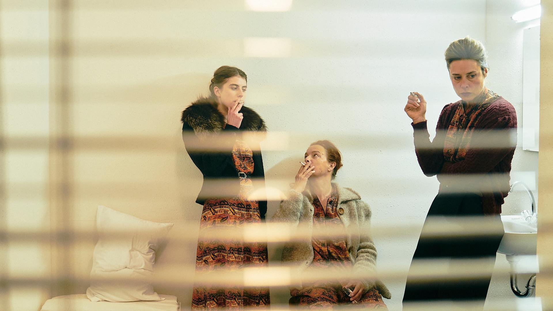 Aufnahme aus einer Szene des Theaterstücks "Berlau :: Königreich der Geister" von "Raum+Zeit" am Berliner Ensemble: Drei Frauen, fotografiert durch ein Fenster hinter geöffneten Jalousien, stehen in einem karg eingerichteten Zimmer und rauchen.