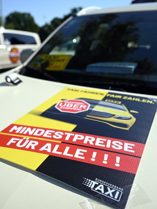 Taxis stehen auf einem Messe-Parkplatz um sich zu einem Autokorso zu sammeln. Auf einer Motorhaube steht auf einem Plakat "Fair zahlen. Mindestpreise für alle"