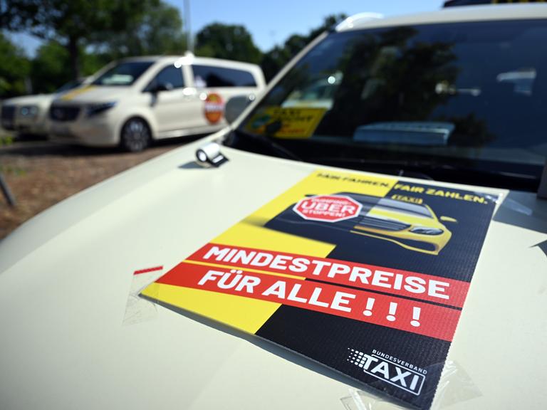 Taxis stehen auf einem Messe-Parkplatz um sich zu einem Autokorso zu sammeln. Auf einer Motorhaube steht auf einem Plakat "Fair zahlen. Mindestpreise für alle"