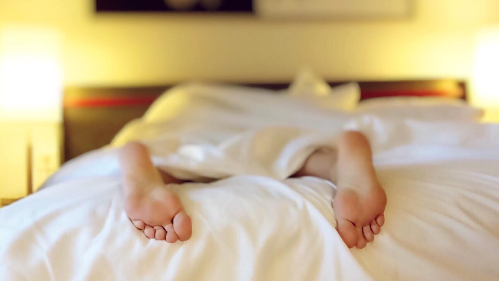 Ein Mann liegt auf dem Bauch in seinem Bett. Man sieht seine Füße unter der Bettdecke hervorschauen.