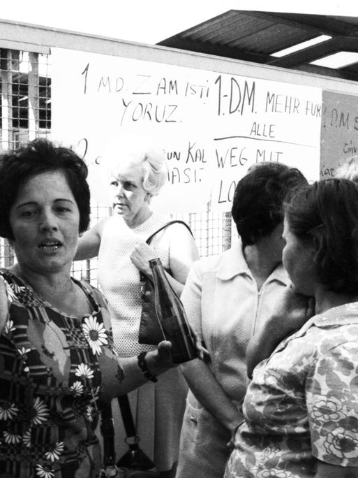 Eine Gruppe von Frauen steht vor einer Tafel, auf der etwas aufgeschrieben ist. Das Bild wirkt, als wäre es im Streikgetümmel entstanden.