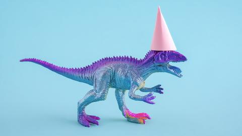 Süßer blau-lila-silbern glitzernder T-Rex mit einem pinken Partyhut auf blauem Hintergrund.