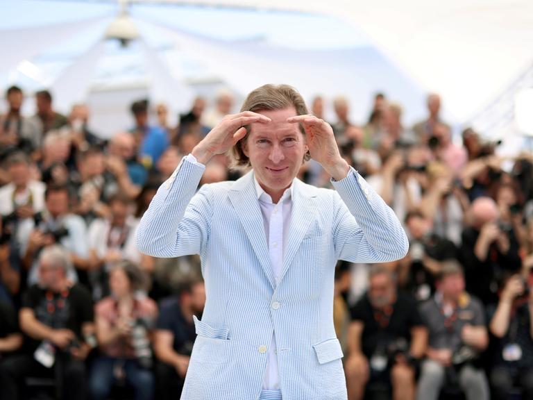 Ein Mann in einem hellblau-weiß gestrieften Anzug hält seine Hände recht und links nehmen die Augen und schaut in Richtung Kamera. Es ist der Regisseur Wes Anderson.