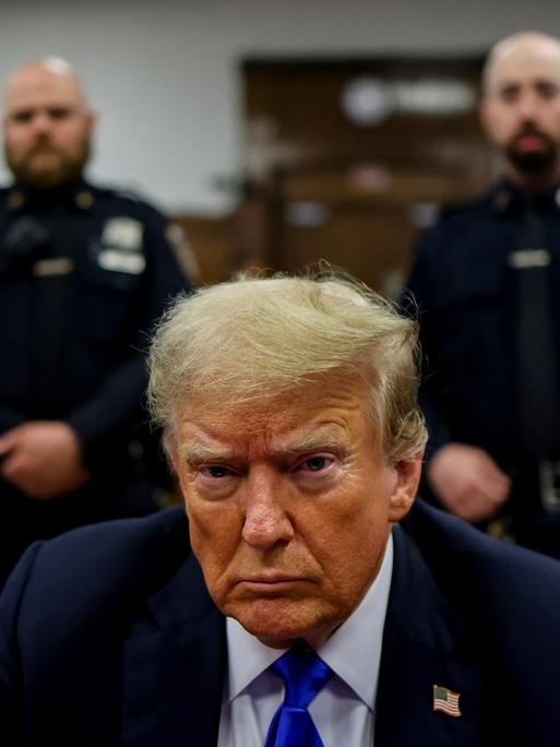 Donald Trump sitzt vor Gericht und schaut ernst in die Kamera. Hinter ihm wachen zwei Polizisten.