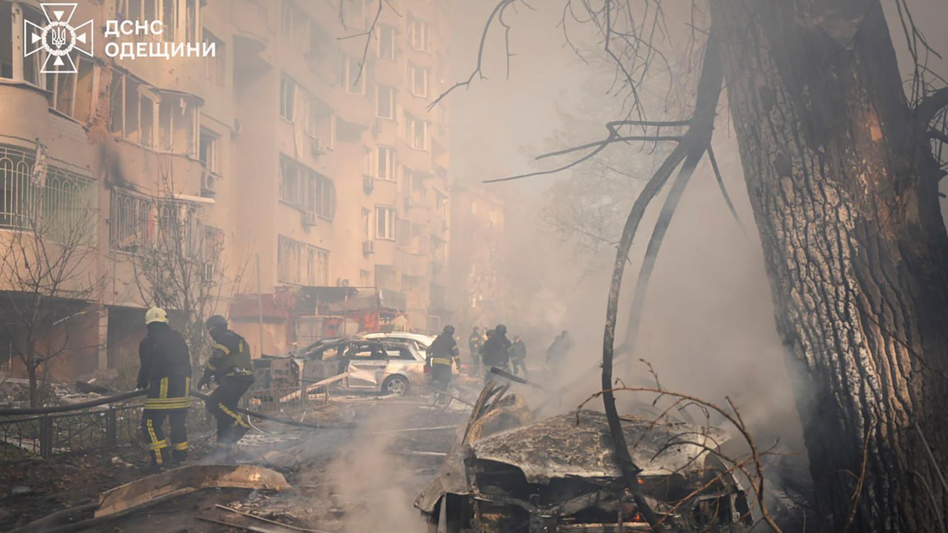 Vor einem zerstörten Wohnblock hängt dichter Rauch. Man erkennt Feuerwehrleute bei Löscharbeiten. 