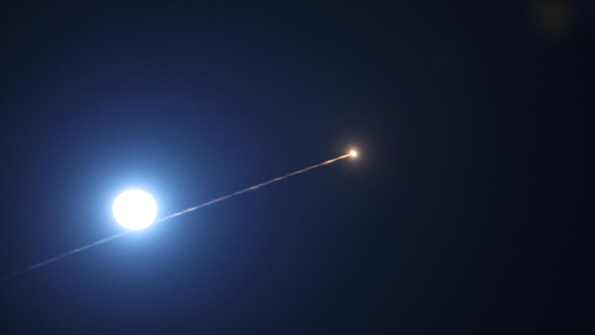 Man sieht den schwarzen Nachthimmel, den weißen Mond und den Leuchtstreifen der Rakete, der quer durchs Bild verläuft. 