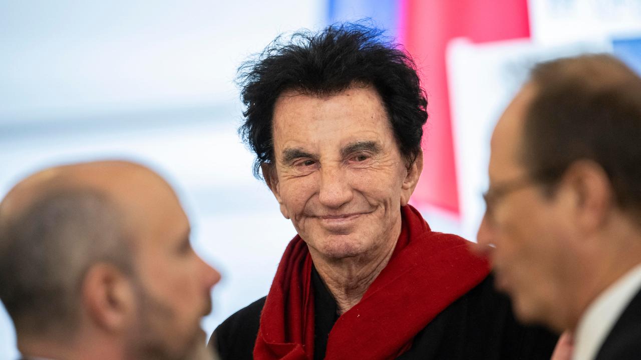 Jack Lang steht zwischen zwei Männern. Er trägt einen roten Schal.