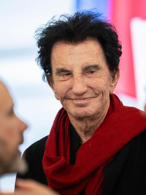 Lang steht lächelnd zwischen zwei Männern. Er trägt einen roten Schal.