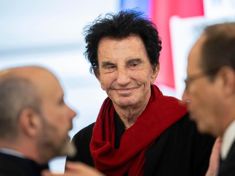 Lang steht lächelnd zwischen zwei Männern. Er trägt einen roten Schal.