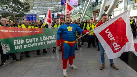 Beschäftigte mit Warnwesten und Transparenten streiken in einer Halle des Flughafens Düsseldorf. Ein Mann im Superhelden-Kostüm hält eine Fahne der Gewerkschaft Verdi.