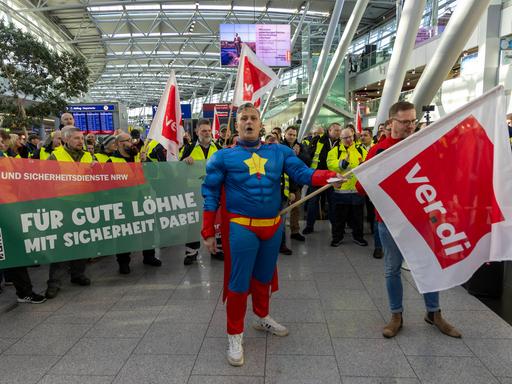 Beschäftigte mit Warnwesten und Transparenten streiken in einer Halle des Flughafens Düsseldorf. Ein Mann im Superhelden-Kostüm hält eine Fahne der Gewerkschaft Verdi.