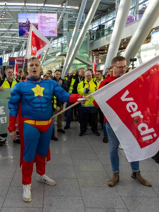 Beschäftigte mit Warnwesten und Transparenten streiken in einer Halle des Flughafens Düsseldorf. Ein Mann im Superhelden-Kostüm hält eine Fahne der Gewerkschaft Verdi.