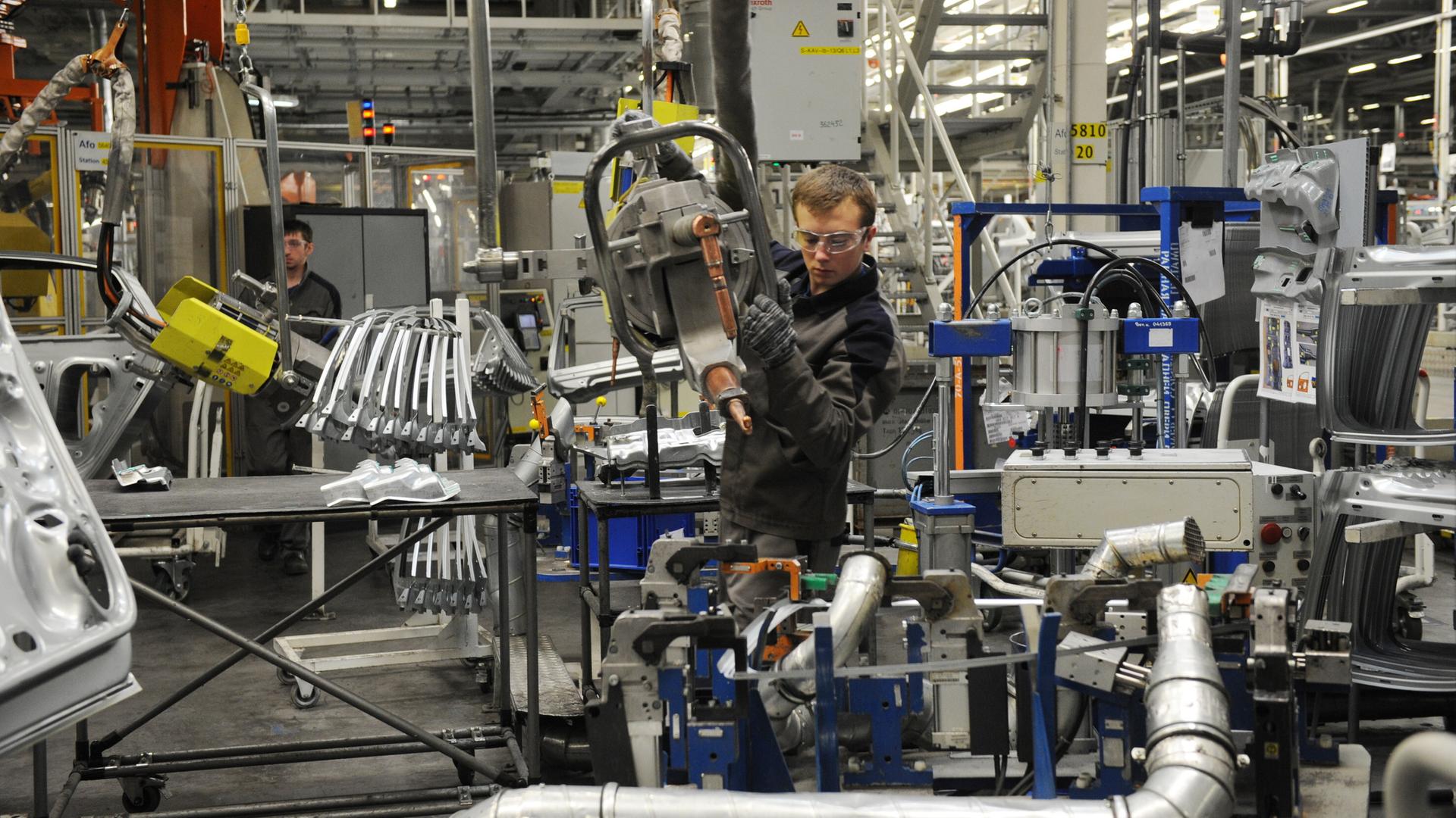 Ein Arbeiter mit Schutzbrille führt ein von der Decke hängendes Gerät an einen VW-Motor. Dahinter steht ein weiterer Arbeiter.