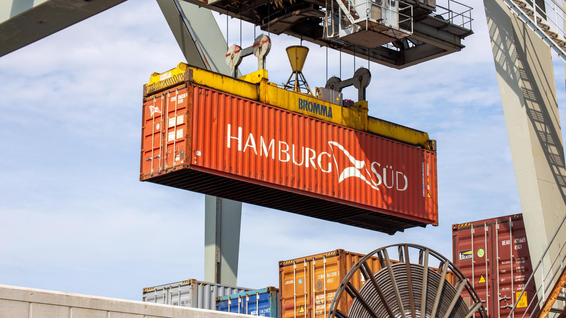 Blick von unten in die Höhe: Ein roter Container mit der Aufschrift "Hamburg-Süd" wird gerade von einem Kran gehoben. 