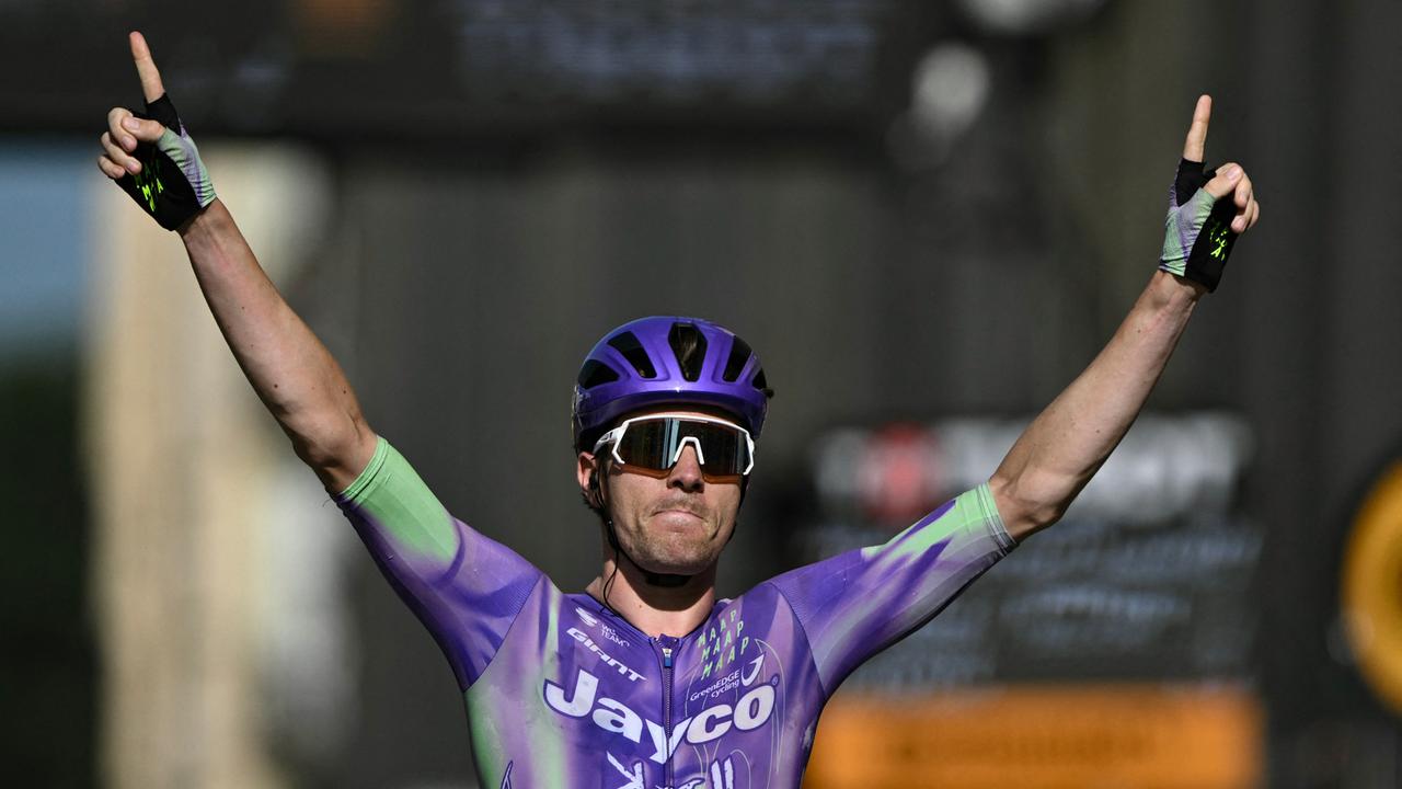 Der Schweizer Radprofi Mauro Schmid im lila Trikot und mit helm und Spoerrtsonnenbrille fährt durch den Zielbereich und streckt beide Arme nach oben. 
