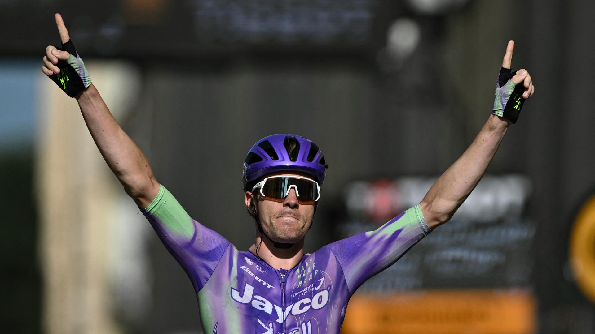 Der Schweizer Radprofi Mauro Schmid im lila Trikot und mit helm und Spoerrtsonnenbrille fährt durch den Zielbereich und streckt beide Arme nach oben. Der Schweizer Radprofi Mauro Schmid im lila Trikot und mit helm und Spoerrtsonnenbrille fährt durch den Zielbereich und streckt beide Arme nach oben.