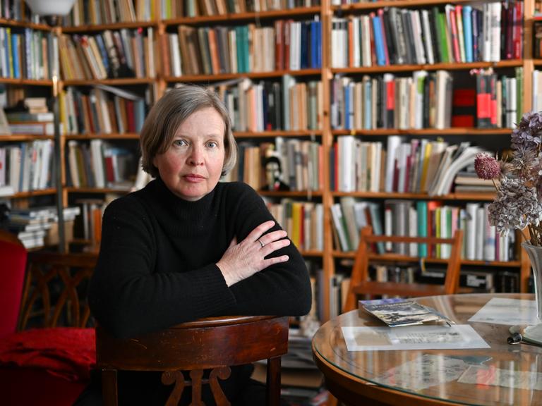 Die Schriftstellerin Jenny Erpenbeck in ihrem Arbeitszimmer. Sie sitzt rittlings auf einem Stuhl vor einer Bücherwand.