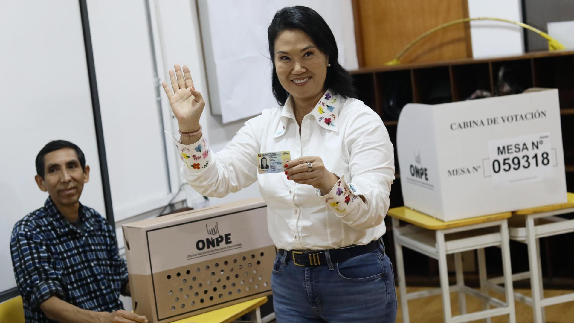 Fujimori im weißen Hemd steht vor einer Wahlurne, hält ihren Personalausweis vor sich und lacht. Sie winkt mit der rechten Hand . Neben ihr sitzt ein Wahlhelfer an einem Tisch. 